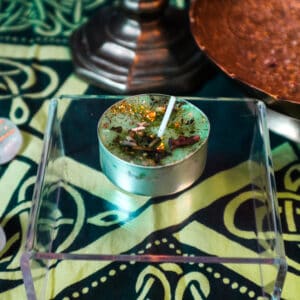 Grüne Teekerze Geldsegen mit goldenem Glitzer und Kräutern auf durchsichtigem Podest mit Runen als Deko