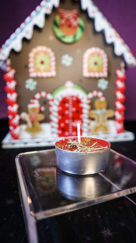 Teekerze Weihnachtswunsch in Rot mit Glitzer und Tannennadeln auf durchsichtigem Podest mit Lebkuchenhaus als Deko im Hintergrund