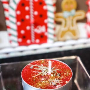 Rote Teekerze Weihnachtswunsch mit Glitzer und Tannennadeln auf durchsichtigem Podest mit Lebkuchenhaus als Deko im Hintergrund