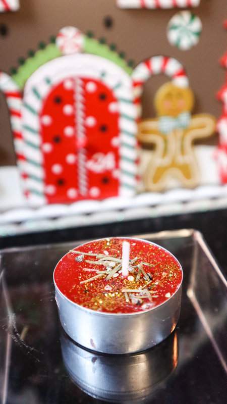 Rote Teekerze Weihnachtswunsch mit Glitzer und Tannennadeln auf durchsichtigem Podest mit Lebkuchenhaus als Deko im Hintergrund
