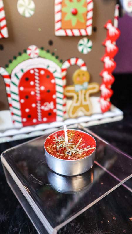 Teekerze Weihnachtswunsch rot mit Glitzer und Tannennadeln auf durchsichtigem Podest mit Lebkuchenhaus im Hintergrund